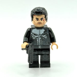 Könnte beinhalten: Eine Kunststoff-Minifigur in schwarzer taktischer Kleidung mit einem weißen Totenkopf-Emblem auf der Brust. Die Figur hat dunkles Haar, ein Gesicht mit Stoppeln und roten Markierungen und hält eine kleine schwarze Pistole in der rechten Hand.