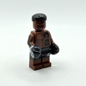 Könnte beinhalten: Eine braune Kunststoff-Minifigur mit schwarzer Frisur, Gesichtstattoos und Brusttattoos. Die Figur trägt schwarze Boxhandschuhe mit weißen Akzenten und eine schwarze Hose mit einem kleinen US-Flaggen-Emblem auf der Vorderseite.