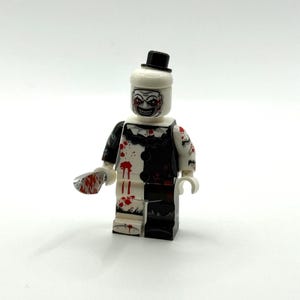 Könnte beinhalten: Eine Kunststoff-Minifigur mit schwarzem Zylinder und einem schwarz-weißen Clownskostüm mit roten Blutspritzern. Die Figur hat ein weißes Gesicht mit einem breiten, zahnigen Grinsen und hält ein kleines silbernes Messer mit roten Blutflecken.