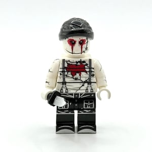 May include: A plastic minifigure featuring a white head with red eye markings and black hair. The torso displays a red chest wound, black suspenders, and barbed wire patterns. The legs are black with white line details, and a small silver knife is held.