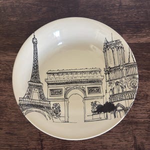 May include: A round, cream-colored ceramic bowl featuring a detailed black line drawing of iconic Parisian landmarks, including the Eiffel Tower, the Arc de Triomphe, and the Notre-Dame Cathedral, arranged in a continuous cityscape design.
