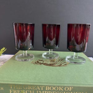May include: Three matching stemmed glasses with deep red, translucent bowls and clear glass bases. The glasses feature a vertical ribbed texture along the sides of the bowls. Each glass has a short, sturdy stem connecting the bowl to the circular base.