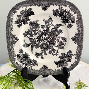 May include: A square ceramic plate featuring a black and white transferware pattern. The design displays intricate floral arrangements and two birds centered on the plate, surrounded by a decorative border with a fine honeycomb-like texture.