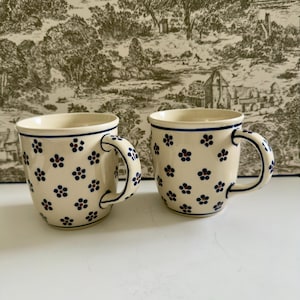 May include: Two cream-colored ceramic mugs featuring a repeating pattern of small blue flowers with dark centers. Each mug has a curved handle and a thin blue line encircling the rim. The mugs are identical in shape, size, and floral design.