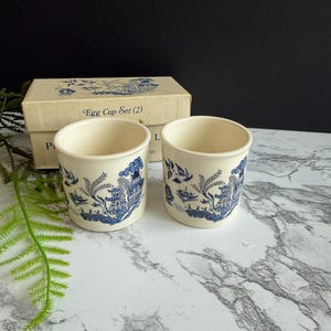 May include: A set of two cream-colored ceramic egg cups featuring a blue willow pattern with pagodas, bridges, and birds. The cups are displayed in front of their original beige cardboard box, which is labeled with the text Egg Cup Set (2).