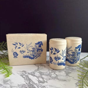 May include: A pair of cylindrical ceramic salt and pepper shakers in an off-white finish, each decorated with a blue willow pattern featuring birds, a boat, and a pagoda. The set is accompanied by a matching rectangular cardboard box.