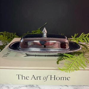 May include: A polished silver-tone metal butter dish with a rectangular base and a matching lid. The lid features a decorative beaded edge and a small, pointed finial handle at the center. The dish has a reflective surface and a classic design.