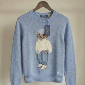 May include: A light blue crewneck sweater featuring a knit graphic of a teddy bear wearing a cap, cream sweater, and blue jeans. The sweater has a ribbed collar, cuffs, and hem, with the letters RL embroidered in white on the lower right side.