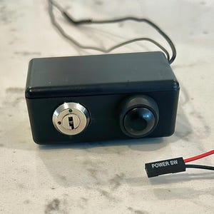 May include: A rectangular black control box featuring a silver key-operated lock on the left and a circular black push button on the right. A thin black wire extends from the top, and a separate red and black wire connector labeled POWER SW is included.