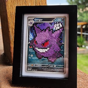 May include: A framed cross-stitch embroidery of the Gengar Pokémon character, featuring a purple creature with red eyes and a white grin, layered over a Gengar trading card with 130 HP, all encased in a simple black rectangular frame.