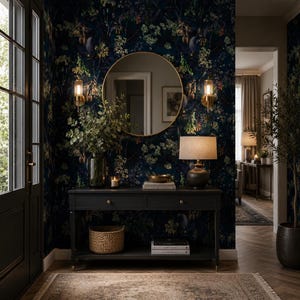 May include: A dark charcoal console table with two drawers and a lower shelf, positioned against a wall with dark floral wallpaper. The table holds a glass vase with greenery, a candle, a bowl, and a lamp, with a round gold mirror above it.