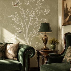 May include: A dark green velvet sofa and armchair with gold patterned cushions, a round wooden side table with a brass lamp, and a green wall featuring a white tree and bird mural. A framed landscape painting hangs on the wall above the armchair.