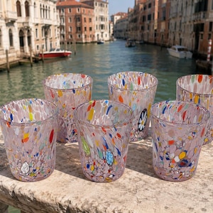 May include: A set of five clear glass tumblers featuring a light pink base colour with scattered, multi-coloured confetti-like flecks in shades of blue, yellow, orange, and white. Each glass has a slightly tapered shape and a smooth, rounded rim.