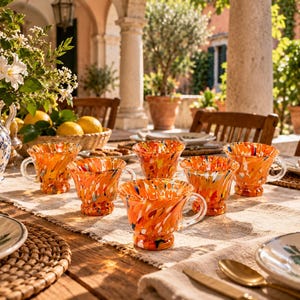 Könnte beinhalten: Ein Set aus fünf Glastassen mit einem leuchtend orangefarbenen, gelben und weißen Sprenkelmuster. Jede Tasse hat einen ausgestellten Rand, einen kurzen Standfuß und einen Henkel aus Klarglas, angeordnet auf einer strukturierten Stoffoberfläche.