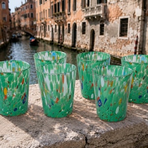 Peut inclure: Un ensemble de cinq verres à boire présentant une couleur de base vert vif avec des éclats multicolores de verre blanc, jaune, orange et bleu dispersés sur toute la surface. Chaque verre a une forme cylindrique à parois droites et un bord lisse.