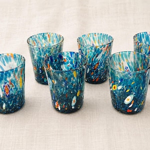 Könnte beinhalten: Ein Set aus fünf Trinkgläsern aus Klarglas mit einem gesprenkelten Muster in Blau-, Weiß-, Orange- und Gelbtönen. Jedes Glas hat eine konisch zulaufende zylindrische Form mit einem glatten Rand und einem Boden, der schmaler als die Öffnung ist.