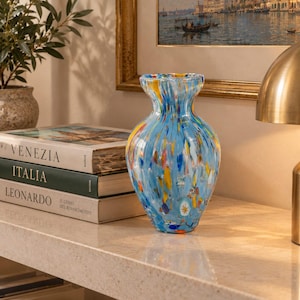 May include: A glass vase with a light blue base featuring a multi-coloured speckled pattern of yellow, orange, white, and dark blue flecks. The vase has a rounded body, a narrow neck, and a flared rim, showcasing a smooth, glossy finish throughout.