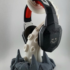 Può includere: Un paio di cuffie da gaming nere con un logo a teschio sul padiglione, poggiate su un supporto bianco a forma di tentacolo di polpo stampato in 3D. Il tentacolo emerge da una base rocciosa grigio scuro e frastagliata che funge da supporto.