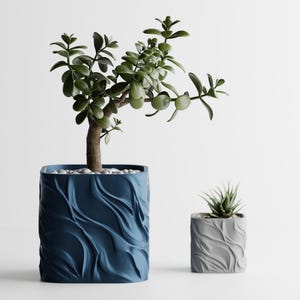 May include: A large dark blue square planter with a textured wavy surface containing a green jade plant and a smaller light gray square planter with a matching wavy texture containing a small succulent, both topped with white decorative pebbles.