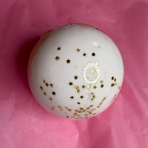 May include: A spherical white bath bomb decorated with numerous small, metallic gold star-shaped sequins scattered across its surface. The object has a smooth, solid texture and a rounded form, featuring a consistent distribution of gold stars.