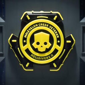May include: A circular black and yellow emblem featuring a central skull icon. The text MALEVELON CREEK VETERAN and HELLDIVERS II is printed in yellow. The emblem has a mechanical design with three protruding side sections.