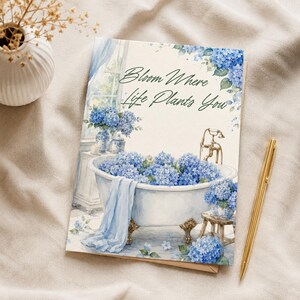 Könnte beinhalten: Eine rechteckige Grußkarte mit einer Aquarellillustration einer weißen Badewanne mit Klauenfüßen, die mit blauen Hortensien gefüllt ist. Der Text Bloom Where Life Plants You ist in dunkler Schreibschrift gedruckt. Ein hellblaues Handtuch hängt über dem Rand.