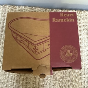 May include: A brown cardboard box for a Heart Ramekin. The box features a maroon side panel with the text Heart Ramekin and a circular logo indicating the item is dishwasher, oven, and freezer safe. A line drawing of the heart-shaped dish is visible.