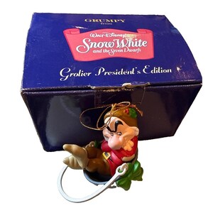 May include: A Grumpy figurine ornament from Snow White and the Seven Dwarfs Grolier President's Edition, featuring the character in a red tunic and brown pants, sitting in a bucket with a white handle, packaged in a dark blue box.