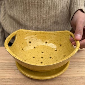 May include: A speckled yellow ceramic colander with two handles and a matching base. The colander has multiple drainage holes. The item is handmade and suitable for kitchen use.