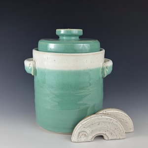 May include: A ceramic fermentation crock with a teal glaze and a white band. The crock has a matching lid and two white, arched weights. The crock has two handles and is designed for pickling or fermenting vegetables.