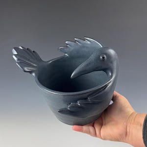 May include: A ceramic bowl in the shape of a bird, with a dark blue-gray glaze. The bowl features a beak, wings, and tail, with the bowl's opening at the bird's back. The bowl is held by a person's hand.