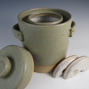 May include: A green ceramic crock with a lid and three white ceramic weights. The crock has a speckled glaze and a beige rim. The lid has a knob on top.