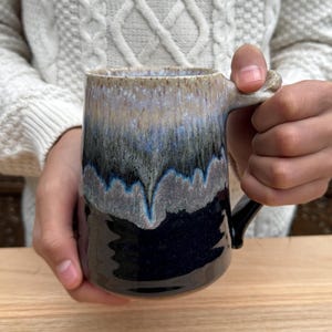Peut inclure: Une tasse en céramique faite à la main avec un glaçage unique. La tasse présente une base bleu marine foncé qui se transforme en gris, bleu et beige en haut. La poignée est noire. La tasse est tenue par une personne.