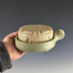 May include: A ceramic butter dish in the shape of a turtle. The lid is tan with a carved geometric design. The base is a light green with small feet. The turtle's head and tail are part of the design.