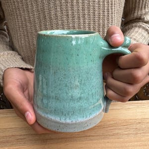 Könnte beinhalten: Eine handgefertigte Keramiktasse mit einer meergrünen Glasur und einem hellgrauen Boden. Die Tasse hat ein einzigartiges Griffdesign und wird mit beiden Händen gehalten. Diese Steinzeugtasse ist perfekt für Kaffee oder Tee.