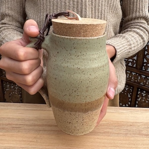 May include: A handmade ceramic mug with a cork lid. The mug features a speckled, two-tone design in green and tan. A leather cord is attached to the lid and handle. The mug is held by a person.