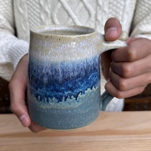 Może przedstawiać: Ręcznie wykonany ceramiczny kubek z gradientem kolorów. Góra jest jasno beżowa, przechodząca w biel, następnie odcienie niebieskiego i kończąca się ciemniejszym szaro-niebieskim u podstawy. Kubek ma solidny uchwyt i teksturowaną powierzchnię.
