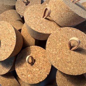 May include: A collection of cork bottle stoppers with a natural, textured appearance. Each stopper has a rounded top and a leather loop handle. The stoppers are light brown and vary slightly in shape and size.