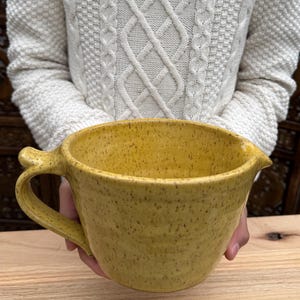 May include: A handmade yellow ceramic mixing bowl with a handle and a pouring spout. The bowl has a speckled texture and a rustic appearance, perfect for kitchen use. The bowl is held in front of a person wearing a white cable knit sweater.