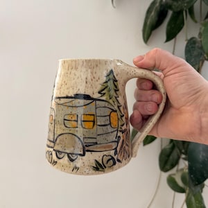 May include: A speckled ceramic mug featuring a hand-painted illustration of a vintage camper trailer parked next to a pine tree and a small campfire. The mug has a cream-colored base with dark brown flecks and a sturdy handle on the right side.