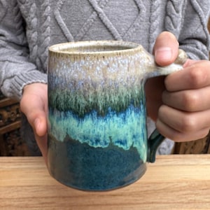 Puede incluir: Una taza de cerámica hecha a mano con un esmalte azul, verde y blanco. La taza tiene un cuerpo ancho y redondeado y una asa.