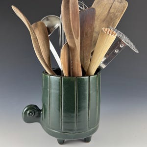 May include: A dark green ceramic utensil holder shaped like a turtle with a textured surface. The holder is filled with various wooden kitchen utensils, including spoons, spatulas, and a whisk.