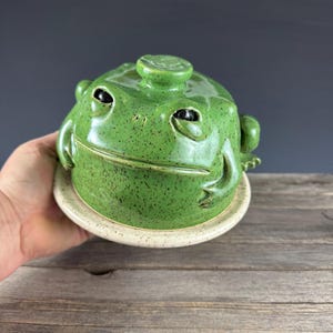 Peut inclure: Un beurrier en céramique en forme de grenouille, avec un glaçage vert tacheté. Le plat a une base ronde et un couvercle avec un petit bouton. Les yeux de la grenouille sont noirs.
