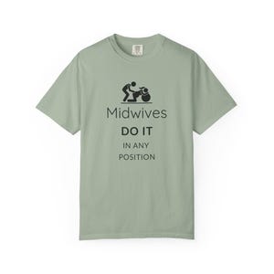 Puede incluir: Una camiseta de manga corta color verde salvia con un gráfico negro de dos figuras junto a una pelota de parto y el texto: Midwives DO IT IN ANY POSITION. La prenda cuenta con cuello redondo y un corte estándar con letras negras.