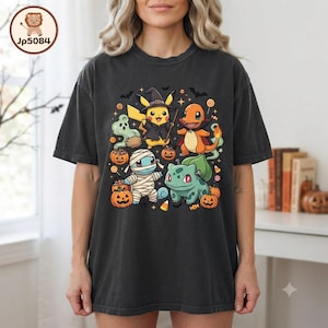 Könnte beinhalten: Ein anthrazitfarbenes Kurzarm-T-Shirt mit einem Grafikdruck von vier Zeichentrickfiguren in Halloween-Kostümen, darunter eine Hexe, ein Vampir, eine Mumie und Frankensteins Monster, umgeben von Kürbissen und Süßigkeiten.