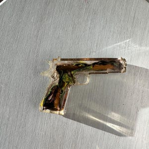 May include: A translucent resin handgun replica featuring a marbled interior design with swirls of dark brown, black, and vibrant green colors. The object has a smooth, glossy finish and a clear outer edge that catches the light, creating a distinct shadow.