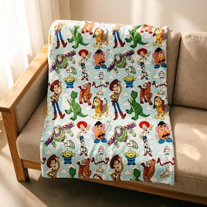 May include: A light blue fleece blanket featuring a repeating pattern of Toy Story characters including Woody, Buzz Lightyear, Jessie, Rex, Slinky Dog, Mr. Potato Head, Forky, and the green aliens, all set against a background of white clouds.