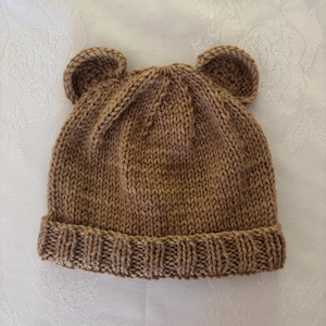 Pode incluir: Um gorro de malha castanho-claro com duas pequenas orelhas arredondadas semelhantes às de um urso no topo. O gorro é construído com um padrão de ponto jersey no corpo principal e uma textura canelada ao longo da aba dobrada.