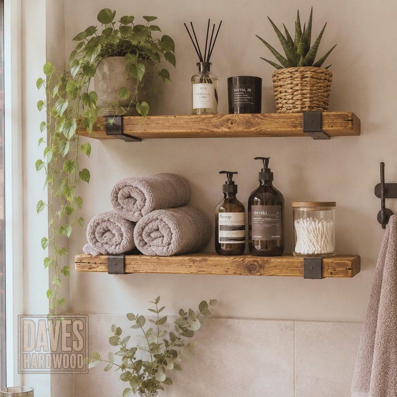 Puede incluir: Dos estantes flotantes de madera r&uacute;stica con soportes de metal negro montados en una pared. El estante superior sostiene una planta verde colgante, un difusor de varillas, una vela negra y una suculenta en una cesta tejida. El estante inferior contiene tres toallas grises enrolladas, dos botellas de vidrio &aacute;mbar con dosificador y un frasco de vidrio lleno de bastoncillos de algod&oacute;n.