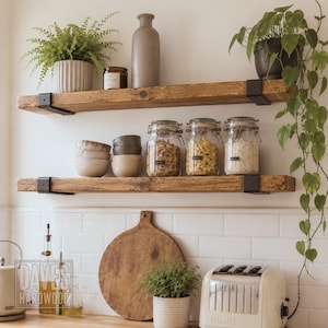 Puede incluir: Dos estantes flotantes de madera rústica con soportes de metal negro montados en una pared de azulejos blancos. Los estantes sostienen cuencos de cerámica, frascos de vidrio con cierres metálicos, un jarrón gris alto, una vela y un helecho.